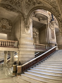 An ornate interior featuring a grand marble staircase with intricately carved stone railings. The architecture includes elaborate decorative elements on the ceiling and walls, with detailed sculptures and lavish molding.