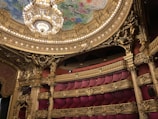 An opulent theater interior featuring grand architectural details with intricate gold ornamentation. A massive chandelier hangs from the ornate ceiling, which displays colorful artwork. The seating area is plush, with rows of luxurious red seats.