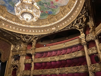 An opulent theater interior featuring grand architectural details with intricate gold ornamentation. A massive chandelier hangs from the ornate ceiling, which displays colorful artwork. The seating area is plush, with rows of luxurious red seats.