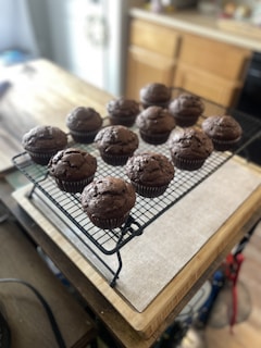 A batch of freshly baked chocolate muffins rests on a cooling rack placed on a wooden cutting board. The muffins have a rich, dark chocolate color with a slightly cracked top, indicating a soft texture inside. The background shows a kitchen setting with cabinets and a countertop, slightly blurred to emphasize the muffins.