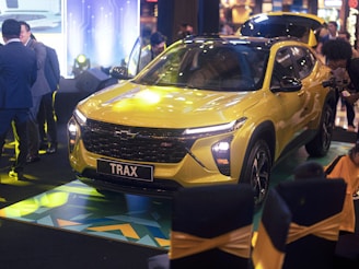 A bright yellow SUV with sleek, modern design features is displayed at an indoor event. The vehicle is surrounded by several people, who are likely attendees at the event. The scene is well-lit with ambient lighting, highlighting the car's glossy finish. In the background, various electronic displays and decor contribute to the event atmosphere.