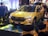 A bright yellow SUV with sleek, modern design features is displayed at an indoor event. The vehicle is surrounded by several people, who are likely attendees at the event. The scene is well-lit with ambient lighting, highlighting the car's glossy finish. In the background, various electronic displays and decor contribute to the event atmosphere.