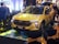 A bright yellow SUV with sleek, modern design features is displayed at an indoor event. The vehicle is surrounded by several people, who are likely attendees at the event. The scene is well-lit with ambient lighting, highlighting the car's glossy finish. In the background, various electronic displays and decor contribute to the event atmosphere.