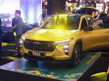 A vibrant yellow car is prominently displayed in an indoor setting with bright lights illuminating its sleek, modern design. Several people in formal attire are nearby, indicating an event or presentation. The background features a digital, futuristic design element complemented by atmospheric lighting.