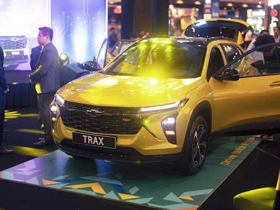 A vibrant yellow car is prominently displayed in an indoor setting with bright lights illuminating its sleek, modern design. Several people in formal attire are nearby, indicating an event or presentation. The background features a digital, futuristic design element complemented by atmospheric lighting.