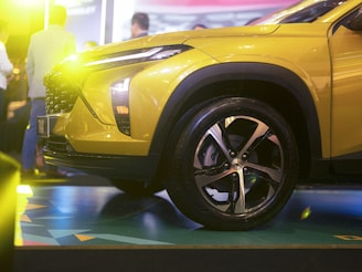 A bright, yellow vehicle is prominently displayed indoors with showroom lighting casting a warm glow. The focus is on the front side of the car, highlighting the wheel, headlight, and aerodynamic design details. Several people in the background are blurred, suggesting a busy event or car show atmosphere.
