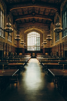 A grand, medieval-style hall with high ceilings adorned with intricate wood carvings and large chandeliers. Tall arched windows allow soft light to filter in, illuminating long wooden tables and chairs arranged in rows across the polished floor. The space exudes a sense of history and elegance.