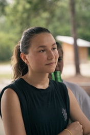 A young person is standing outdoors with greenery in the background. They have a contemplative expression and are wearing a black sleeveless shirt. A faint image of another person can be seen in the background, partially obscured.