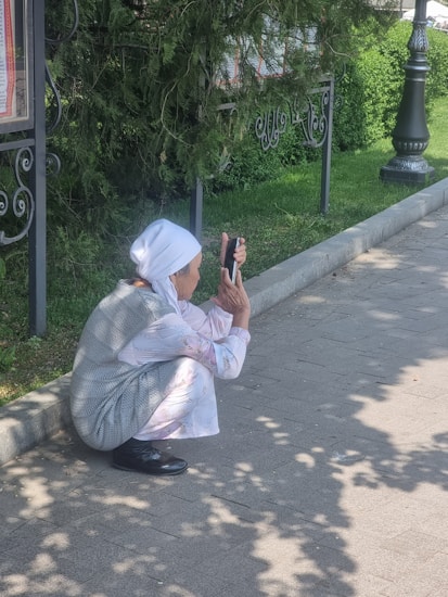 An elderly person wearing a white headscarf and light clothing is squatting on a sidewalk, using a smartphone. They are near a grassy area with a hedge and wrought iron fencing, partially shaded by trees.