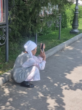 An elderly person wearing a white headscarf and light clothing is squatting on a sidewalk, using a smartphone. They are near a grassy area with a hedge and wrought iron fencing, partially shaded by trees.