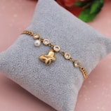 A gold bracelet with a butterfly charm and multiple round, clear gemstones is displayed on a soft, gray cushion. There is also a single pearl detail attached to the bracelet. The background has a soft pink hue with a hint of blurred floral elements.