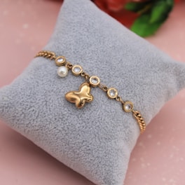 A gold bracelet with a butterfly charm and multiple round, clear gemstones is displayed on a soft, gray cushion. There is also a single pearl detail attached to the bracelet. The background has a soft pink hue with a hint of blurred floral elements.