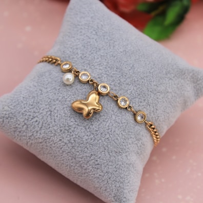A gold bracelet with a butterfly charm and multiple round, clear gemstones is displayed on a soft, gray cushion. There is also a single pearl detail attached to the bracelet. The background has a soft pink hue with a hint of blurred floral elements.