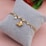 A gold bracelet with a butterfly charm and multiple round, clear gemstones is displayed on a soft, gray cushion. There is also a single pearl detail attached to the bracelet. The background has a soft pink hue with a hint of blurred floral elements.