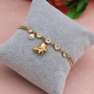 A gold bracelet with a butterfly charm and multiple round, clear gemstones is displayed on a soft, gray cushion. There is also a single pearl detail attached to the bracelet. The background has a soft pink hue with a hint of blurred floral elements.