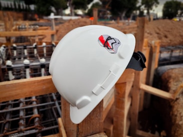 A white construction helmet with a sticker of a character wearing a helmet is resting on a wooden beam at an active construction site. In the background, construction materials and wooden framework are visible, suggesting a building or infrastructure project in progress.