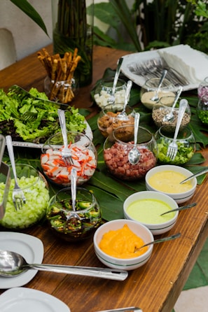 An assortment of fresh ingredients displayed on a wooden table with green leaves as decoration. Glass bowls contain chopped vegetables such as cucumbers, jalapeños, crab sticks, and smoked meats, along with various sauces in smaller bowls. Cutlery is visible alongside stacks of napkins and breadsticks in a separate container.
