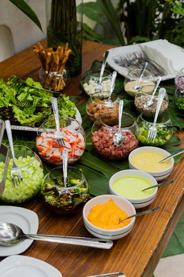 An assortment of fresh ingredients displayed on a wooden table with green leaves as decoration. Glass bowls contain chopped vegetables such as cucumbers, jalapeños, crab sticks, and smoked meats, along with various sauces in smaller bowls. Cutlery is visible alongside stacks of napkins and breadsticks in a separate container.