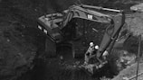 An excavator with the brand name visible on its arm is positioned in a large, earthy excavation site. Two individuals wearing hard hats are seated on the machine’s arm. The site is filled with dark soil, and the scene is monochromatic, giving it a gritty, industrial appearance.