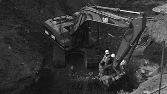 An excavator with the brand name visible on its arm is positioned in a large, earthy excavation site. Two individuals wearing hard hats are seated on the machine’s arm. The site is filled with dark soil, and the scene is monochromatic, giving it a gritty, industrial appearance.