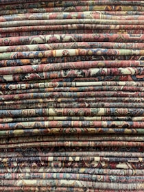 A stack of intricately patterned, colorful woven rugs is displayed, with varying designs and a rich tapestry of colors and textures.