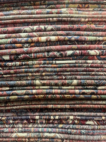 A stack of intricately patterned, colorful woven rugs is displayed, with varying designs and a rich tapestry of colors and textures.