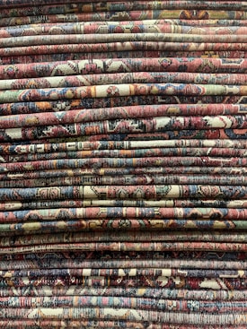 A stack of intricately patterned, colorful woven rugs is displayed, with varying designs and a rich tapestry of colors and textures.