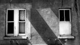 Two old windows on a rough concrete wall, with visible cracks and shadows creating a stark contrast. The left window has broken glass panes and a netted pattern behind it, while the right window is white and appears boarded up.
