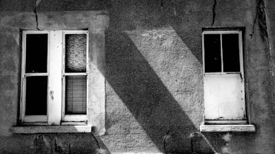 Two old windows on a rough concrete wall, with visible cracks and shadows creating a stark contrast. The left window has broken glass panes and a netted pattern behind it, while the right window is white and appears boarded up.