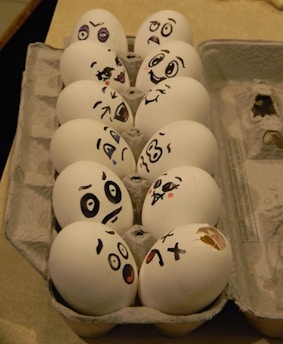 A carton of eggs, each with a playful face drawn in black marker, displaying a variety of exaggerated expressions such as happiness, surprise, confusion, and sadness.