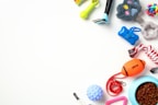 An assortment of pet toys and accessories arranged on a white background. Items include a rope chew toy, a blue feeding bowl filled with kibble, an orange football-shaped toy, a green spiky toy, a small rubber ball, a pair of grooming tools, and a colorful rubber knot ball.
