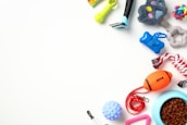 An assortment of pet toys and accessories arranged on a white background. Items include a rope chew toy, a blue feeding bowl filled with kibble, an orange football-shaped toy, a green spiky toy, a small rubber ball, a pair of grooming tools, and a colorful rubber knot ball.