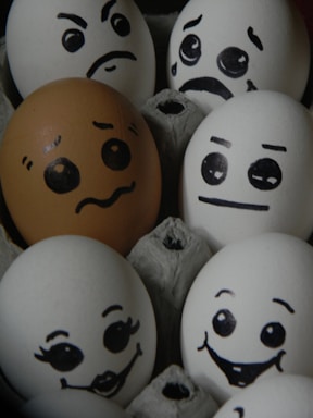 A collection of eggs features hand-drawn expressive faces, each displaying different emotions. The eggs are arranged closely together in an egg carton. One egg is brown with a sad face, while the others are white with happy, angry, or surprised expressions.