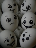 A collection of eggs in a carton, each featuring hand-drawn expressive faces with various emotions, including happiness, surprise, slyness, and curiosity.