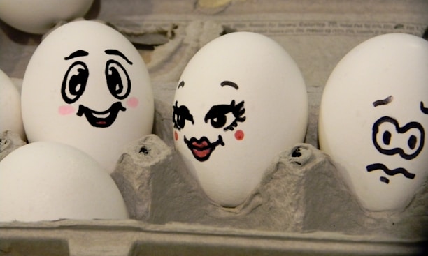 Three eggs in a carton have playful cartoon faces drawn on them using black marker. The first egg has a smiling expression with bright eyes and blushing cheeks. The second egg is decorated with long eyelashes, full lips, and raised eyebrows, giving it a flirty expression. The third egg appears anxious with squiggly eyebrows and a wavy mouth.