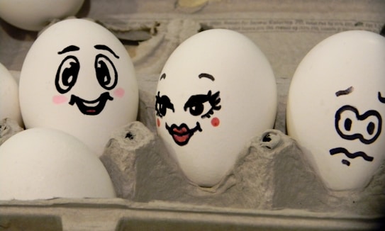 Three eggs in a carton have playful cartoon faces drawn on them using black marker. The first egg has a smiling expression with bright eyes and blushing cheeks. The second egg is decorated with long eyelashes, full lips, and raised eyebrows, giving it a flirty expression. The third egg appears anxious with squiggly eyebrows and a wavy mouth.