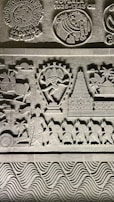 Intricate stone carvings featuring historical symbols and figures, including a dancer with multiple arms, a temple, and a procession of soldiers with shields. Symbols of ancient Indian empires such as Kadamba and Hoysala are prominently displayed along with detailed patterns and artistic elements.