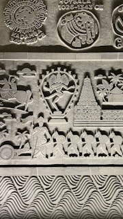 Intricate stone carvings featuring historical symbols and figures, including a dancer with multiple arms, a temple, and a procession of soldiers with shields. Symbols of ancient Indian empires such as Kadamba and Hoysala are prominently displayed along with detailed patterns and artistic elements.