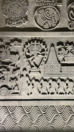 Intricate stone carvings featuring historical symbols and figures, including a dancer with multiple arms, a temple, and a procession of soldiers with shields. Symbols of ancient Indian empires such as Kadamba and Hoysala are prominently displayed along with detailed patterns and artistic elements.