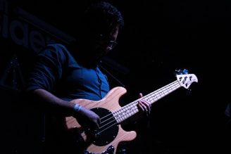 A person is playing an electric bass guitar on a dimly lit stage. The focus is on the wooden body of the guitar, with the strings and tuning pegs visible. The person is wearing glasses and a dark shirt, and the atmosphere is moody with shadows.