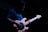 A person is playing an electric bass guitar on a dimly lit stage. The focus is on the wooden body of the guitar, with the strings and tuning pegs visible. The person is wearing glasses and a dark shirt, and the atmosphere is moody with shadows.