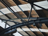 A metal structure with beams and girders supporting a corrugated metal roof. The perspective emphasizes the geometric patterns and lines formed by the overlapping materials.
