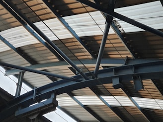 A metal structure with beams and girders supporting a corrugated metal roof. The perspective emphasizes the geometric patterns and lines formed by the overlapping materials.