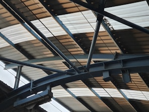 A metal structure with beams and girders supporting a corrugated metal roof. The perspective emphasizes the geometric patterns and lines formed by the overlapping materials.