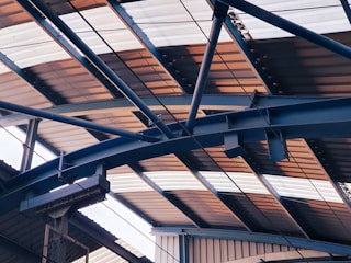 Overhead view of a metal structure with a series of intersecting beams and trusses supporting a corrugated roof, which is partially illuminated by natural light.