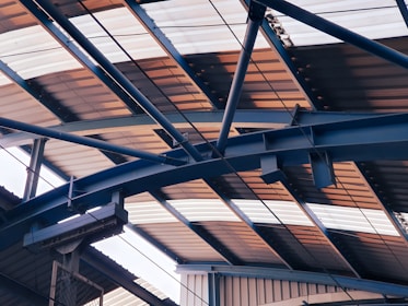 Overhead view of a metal structure with a series of intersecting beams and trusses supporting a corrugated roof, which is partially illuminated by natural light.