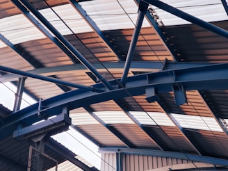 Overhead view of a metal structure with a series of intersecting beams and trusses supporting a corrugated roof, which is partially illuminated by natural light.