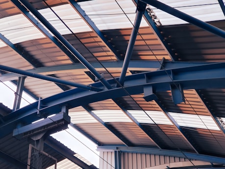 Overhead view of a metal structure with a series of intersecting beams and trusses supporting a corrugated roof, which is partially illuminated by natural light.