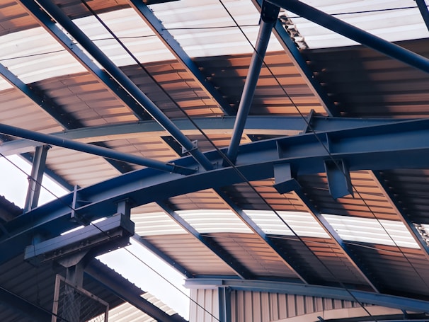 Overhead view of a metal structure with a series of intersecting beams and trusses supporting a corrugated roof, which is partially illuminated by natural light.