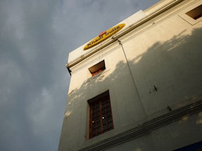 A low-angle view of a beige building with a sign at the top reading 'Khadi India'. The facade features square windows with red frames, and there are shadows cast on the wall, suggesting nearby trees or other structures. The sky is overcast, giving the scene a subdued lighting.